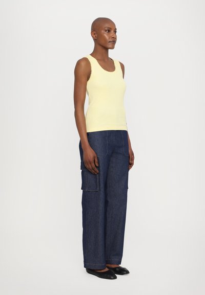 Gelbes, geripptes Tanktop mit Wellenkanten, kombiniert mit dunkelblauen Denim-Cargo-Hosen mit Seitentaschen und Kontrastnähten. Schwarze Ballerinas.