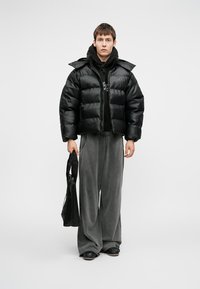 Črna puhovka z sijajno površino, skrajšano obliko in visokim ovratnikom, v kombinaciji z ohlapnimi sivimi hlačami in črnimi torbami.