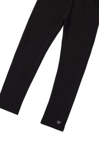 Leggings negros de tela elástica, con un corte ajustado y un pequeño detalle en forma de corazón con strass en la parte inferior izquierda.