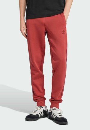 Red jogging pants with elastic cuffs, side pockets, and subtle brand logo. Paired with black and white athletic shoes on a light background.
