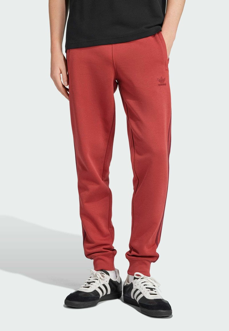 Pantalones deportivos rojos con puños elásticos, bolsillos laterales y un logo de la marca sutil. Combinados con zapatos deportivos en blanco y negro sobre un fondo claro.