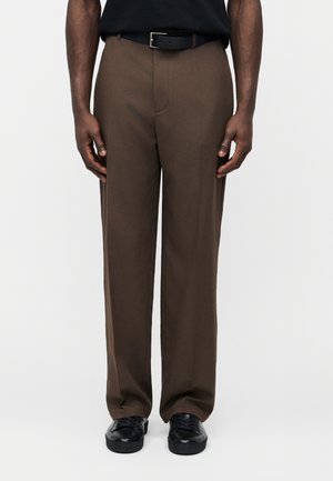 Homme portant un pantalon marron droit, une ceinture noire, une chemise noire et des chaussures noires, debout devant un fond clair uni.