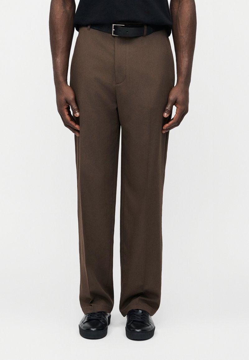 Άνδρας που φοράει καφέ παντελόνι με ίσια γραμμή, μαύρη ζώνη, μαύρο πουκάμισο και μαύρα παπούτσια, στέκεται σε ένα απλό ανοιχτόχρωμο φόντο.