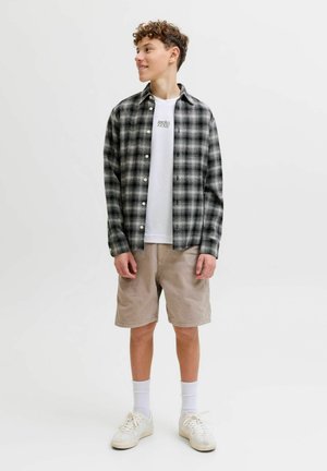 Adolescent debout, portant une chemise à carreaux noir et blanc sur un t-shirt blanc avec logo, un short beige, des chaussettes blanches et des baskets blanches, regardant vers la droite.