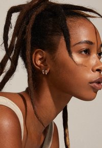 Pendientes de aro de plata adornan la oreja, destacando un perfil lateral con piel texturizada y una camiseta sin mangas neutral frente a un fondo plano.