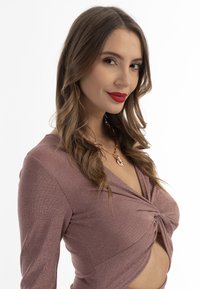 Zawiązany krótki top w różowo-złotym błyszczącym materiale, z długimi rękawami i głębokim dekoltem w serek. Naszyjnik z okrąłym wisiorkiem.