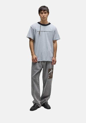 Jeune homme aux cheveux courts et foncés avec une longue tresse, portant une chemise bleu clair avec un texte, un jean gris ample avec une pièce graphique et des chaussures noires.