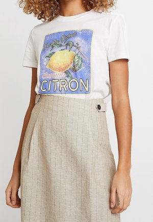 Persoon met krullend haar die een wit T-shirt draagt met een citroengrafiek en tekst "Freshly Picked Summer Citron" en een beige rok met knopen in de taille.
