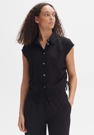 Donna con capelli ricci che indossa una camicia nera senza maniche con bottoni e pantaloni neri, guarda verso sinistra con una mano in tasca.