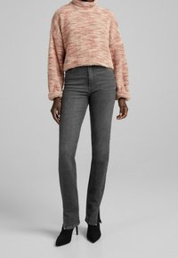 Jasnoróżowy sweter o fakturowanej strukturze z wysokim kołnierzem i luźnym krojem, zestawiony z ciemnoszarymi, wysokim stanem jeansami z rozcięciami przy nogawkach oraz czarnymi szpilkami.