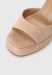 Nude suede korkokenkä, jossa on neliön muotoinen kärki, leveä etuhihna ja teksturoitu pinta varressa ja pohjassa.