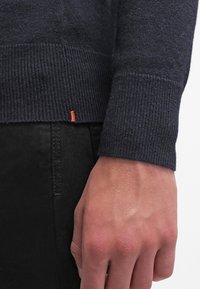 Granatowy sweter z prążkowanymi mankietami, wykonany z teksturowanej tkaniny, z małym pomarańczowym akcentem u dołu. Nosi się go na ciemnych spodniach.