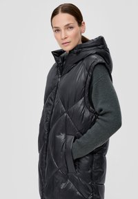 Schwarze Steppweste mit Kapuze, ausgestattet mit einem gesteppte Design, Reißverschluss und Seitentaschen. Getragen über einem dunkelgrünen, strukturierten Pullover.