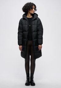 Černá prošívaná péřová bunda s kapucí, sahající do kolen, s prošitým vzorem, předními knoflíky a bočními kapsami, nošená přes tmavé legíny a kozačky.