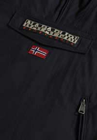 Marinblå jacka med tygstruktur, prydd med en stor broderad logotyp, en liten norsk flagga patch och en ficka med dragkedja på sidan.