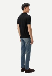 Homme debout, face à droite, portant un polo noir à manches courtes, un jean bleu clair et des chaussures marron foncé sur un fond blanc uni.