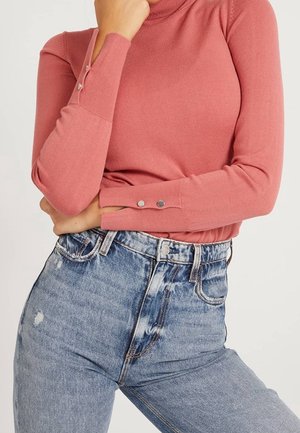 Femme portant un haut à manches longues rose poudré avec des boutons-pression aux poignets et un jean bleu clair délavé taille haute.