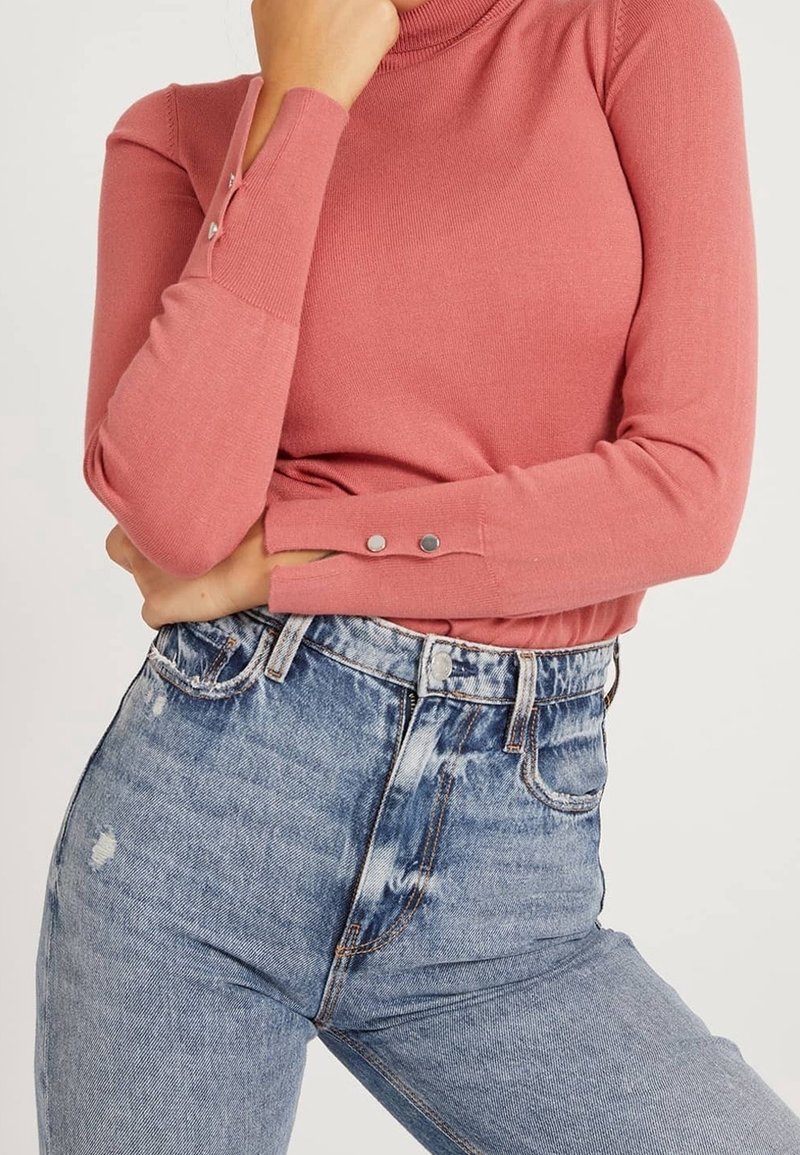 Femme portant un haut à manches longues rose poudré avec des boutons-pression aux poignets et un jean bleu clair délavé taille haute.