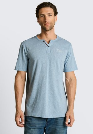 Junger Mann mit kurzen lockigen Haaren, der ein hellblaues Henley-Shirt mit kurzen Ärmeln und blaue Jeans trägt, vor einem schlichten Hintergrund stehend.