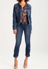 Mörkblå denimjacka med knappstängning, kombinerad med mönstrad blus och mörkblå skinny jeans, med fransade ankelslut och nude-hälar.