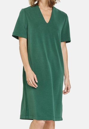 Femme portant une robe verte à manches courtes, longueur genoux, avec un décolleté en V, debout devant un fond clair uni.