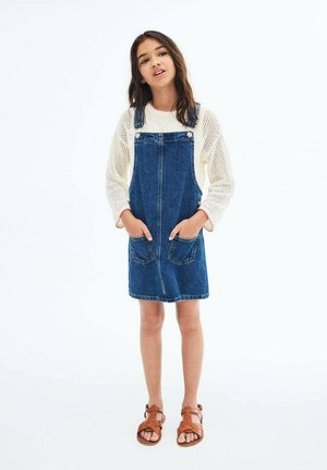 Denim overall jurk, donkerblauw, kniehoogte, met verstelbare banden, twee voorke pockets, gedragen over een crèmekleurige gebreide trui, gecombineerd met bruine sandalen.