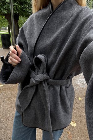 Cappotto avvolgente grigio in lana con maniche larghe, legato in vita, con rifiniture nere. Indossato sopra jeans azzurri chiari, mentre tiene un ombrello.