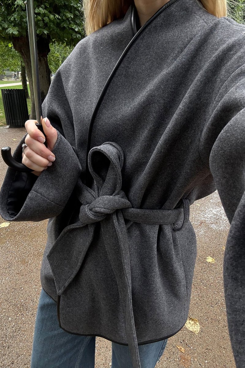 Grå uld indpakningsfrakke med brede ærmer, bundet i taljen, med sort kant. Båret over lyseblå jeans og holder en paraply.