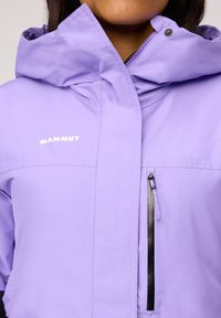 Violetinis vandeniui atsparus švarkas su kapišonu, turintis priekinį užtrauktuką ir krūtinės kišenę su juodu užtrauktuku. Logo kairėje pusėje baltos spalvos.