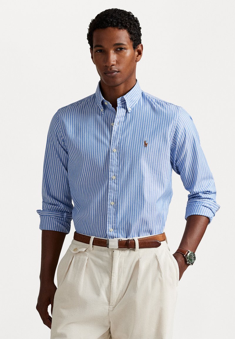 Chemise à boutons rayée bleu clair et blanche avec des manches longues, ornée d'un petit logo sur la poitrine et assortie à un pantalon plissé crème.