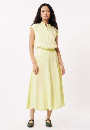 Robe sans manches jaune clair avec un col à boutons et une taille cintrée, descendant jusqu'à une jupe évasée. Chaussures noires et vertes chunky.