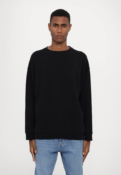Sweatshirt noir avec un col rond, des épaules abaissées et des poignets côtelés. Tissu doux, coupe décontractée, associé à un jean bleu clair.