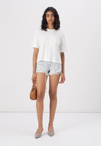 Witte korte mouwen cropped t-shirt met bloemenborduursel, gecombineerd met verstoorde denim shorts. Model houdt een kleine bruine handtas vast.