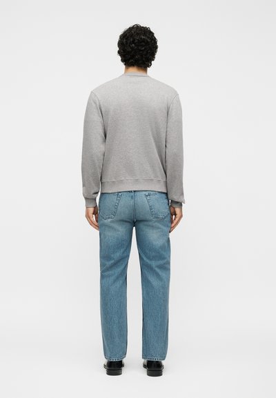 Άνδρας με σγουρά σκούρα μαλλιά, που φοράει γκρι μπλούζα, μπλε τζιν και μαύρα παπούτσια, στέκεται με πλάτη σε απλό λευκό φόντο.