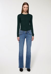 Ciemnozielona bluzka z długim rękawem wykonana z miękkiego materiału, w połączeniu z niebieskimi dzwonami jeansami z neutralnym guzikiem i naszytymi kieszeniami.