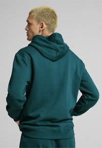 Felpa verde con cappuccio, realizzata in un tessuto strutturato, con polsini e orlo a coste. Presenta un cappuccio grande e una vestibilità morbida, vista dal retro.