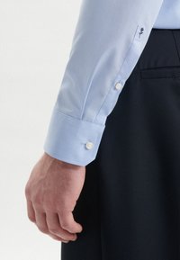 Lichtblauw overhemd met knoopmanchetten en geborduurde bloemdecoratie. De stof is glad en licht glanzend, en sluit nauw aan rond de pols.