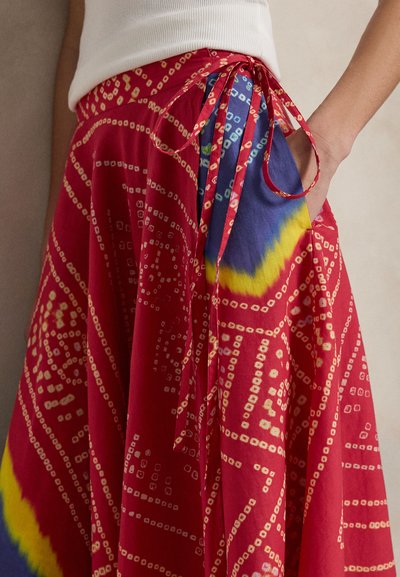 Punainen kietaisuhame, jossa valkoisia geometrisia ja keltasinisiä batikkikuvioita, solmittu vyötärölle, yhdistetty valkoiseen hihattomaan toppiin, käsi taskussa.