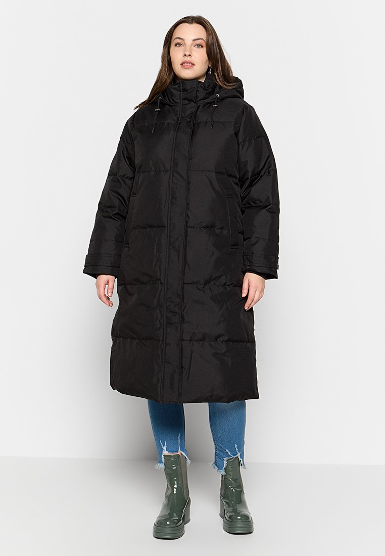 Manteau doudoune noir avec capuche, présentant une coupe longue, un design matelassé et une fermeture éclair à l'avant. Porté sur un jean bleu et des bottines vertes.