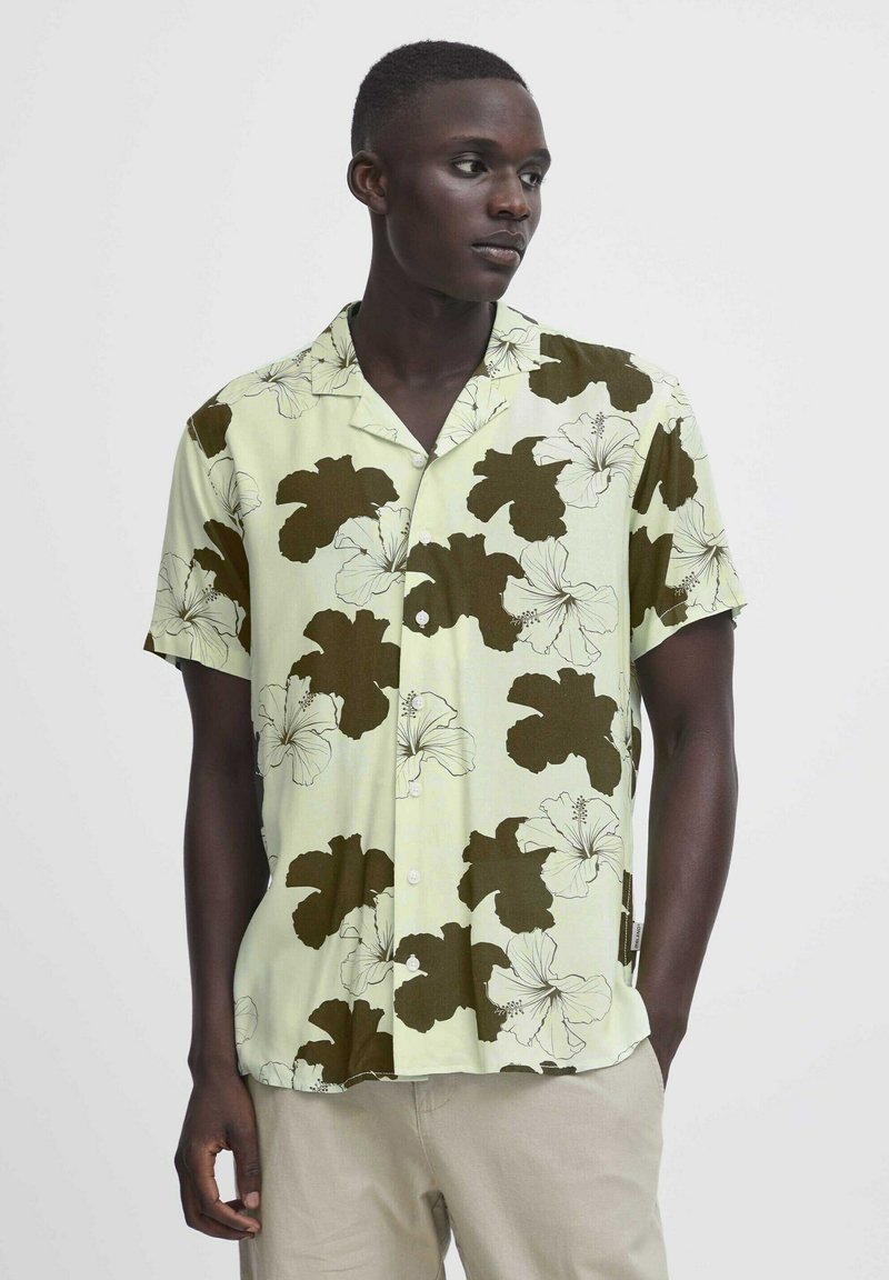 Camisa de manga corta en tela verde claro con un gran patrón floral de hibisco marrón oscuro, cierre de botones y diseño de corte relajado.