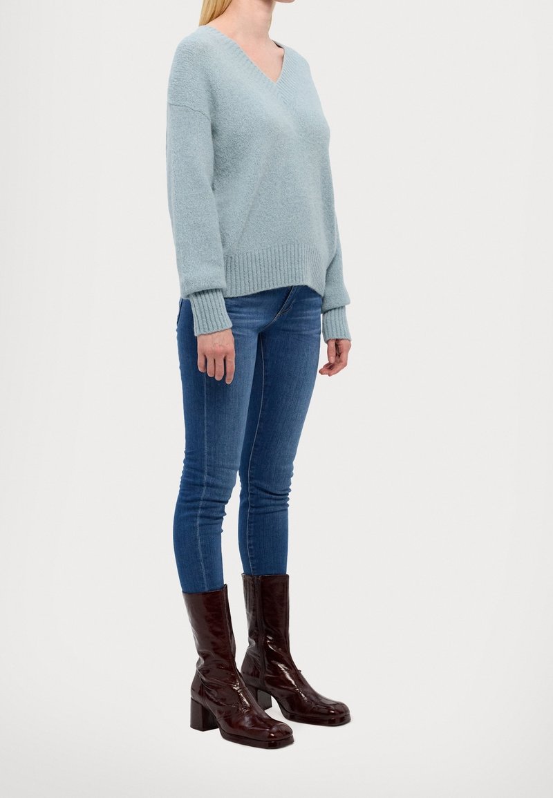 Helles blaues, weiches Strickoberteil mit V-Ausschnitt und gerippten Bündchen, kombiniert mit eng anliegenden blauen Jeans und glänzenden bordeauxfarbenen Stiefeln mit Absatz bis zur Wade.