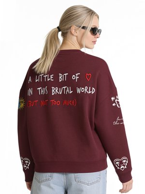 Femme avec une queue de cheval blonde et des lunettes de soleil portant un sweat-shirt bordeaux avec du texte et des dessins colorés de style manuscrit dans le dos et sur les manches.