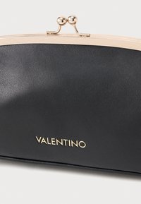 Melnā ādas somiņa ar zelta toni aizdari, kurai ir gluda tekstūra un reljefā "VALENTINO" logo priekšpusē.