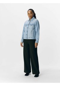 Giacca di jeans azzurra con colletto, tasche frontali e chiusura con bottoni, abbinata a pantaloni neri a gamba larga e tacchi neri.