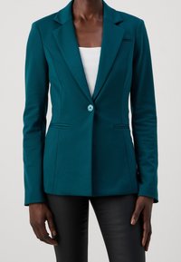 Teal färgad skräddarsydd kavaj med en knapp över en vit ribbad topp, kombinerad med svarta smala byxor, som bärs av en person som står mot en grå bakgrund.