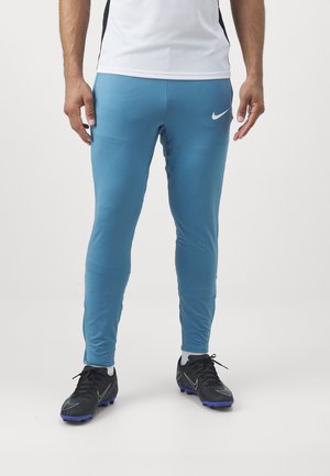 Homme portant un pantalon de sport Nike bleu clair, des chaussettes blanches, des crampons noirs, et une chemise blanche, debout devant un fond uni.