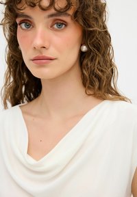 Weiße, drapierte Bluse aus einem weichen, fließenden Stoff. Das Model hat lockige Haare und trägt große Perlenohrringe, die glatte Haut und einen schönen Hals zeigen.