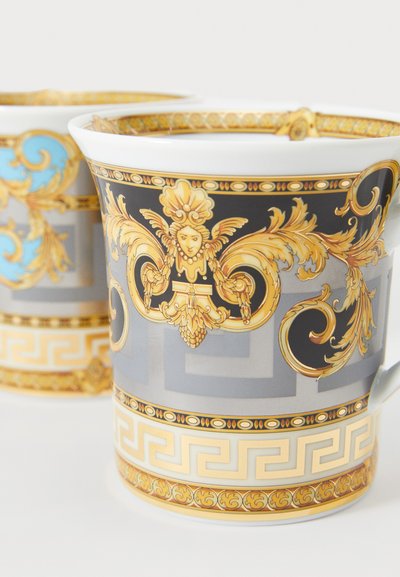 Porzellantasse mit goldenen, verzierten Details auf grauem Hintergrund mit geometrischem Muster. Akzente in Blau und filigrane Designs.