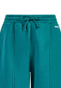 Turkusowe dresy z wiązanym pasem i pionowymi szwami, z małą prostokątną metką marki po prawej stronie.