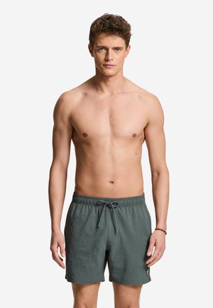 Giovane uomo con capelli ricci che indossa pantaloncini da bagno grigi strutturati con coulisse, in piedi davanti a uno sfondo bianco semplice.